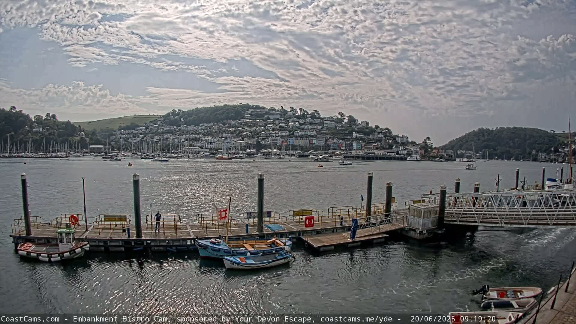 A partly cloudy day shows a harbor with a town on a hill in the background, boats moored at a dock, and smaller boats at a floating pier.