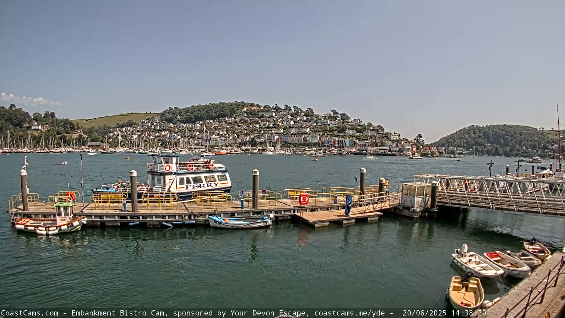 A ferry is docked at a pier in a harbor, with numerous boats and a town on a hillside visible in the background under sunny skies.