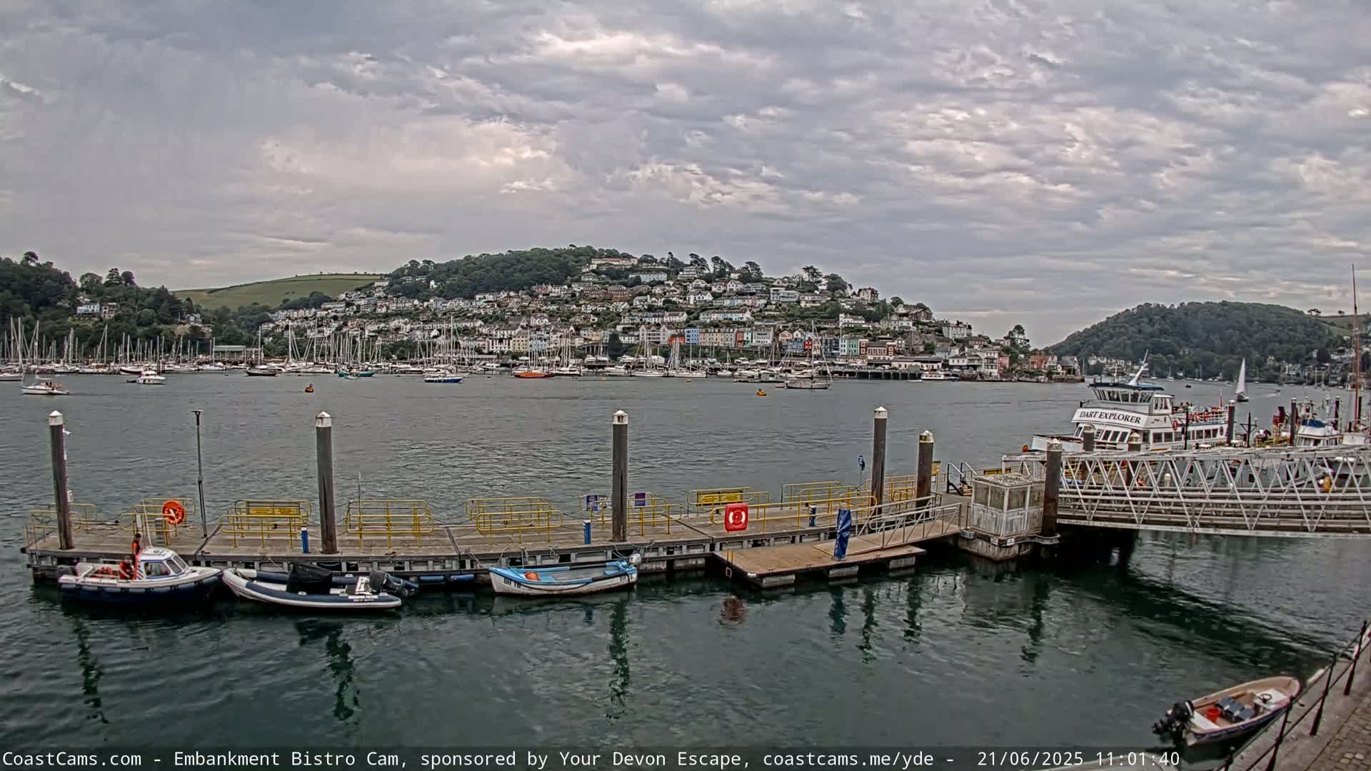 A harbor scene under a cloudy sky shows a town nestled on a hillside overlooking a calm body of water filled with boats and a ferry docked at a pier.
