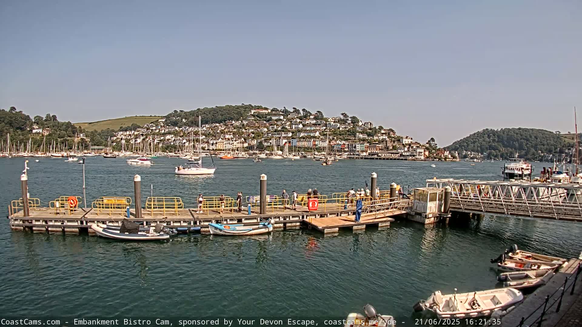 A sunny day at a harbor shows a dock with people on it, many boats in the water, and a town on a hill in the background.