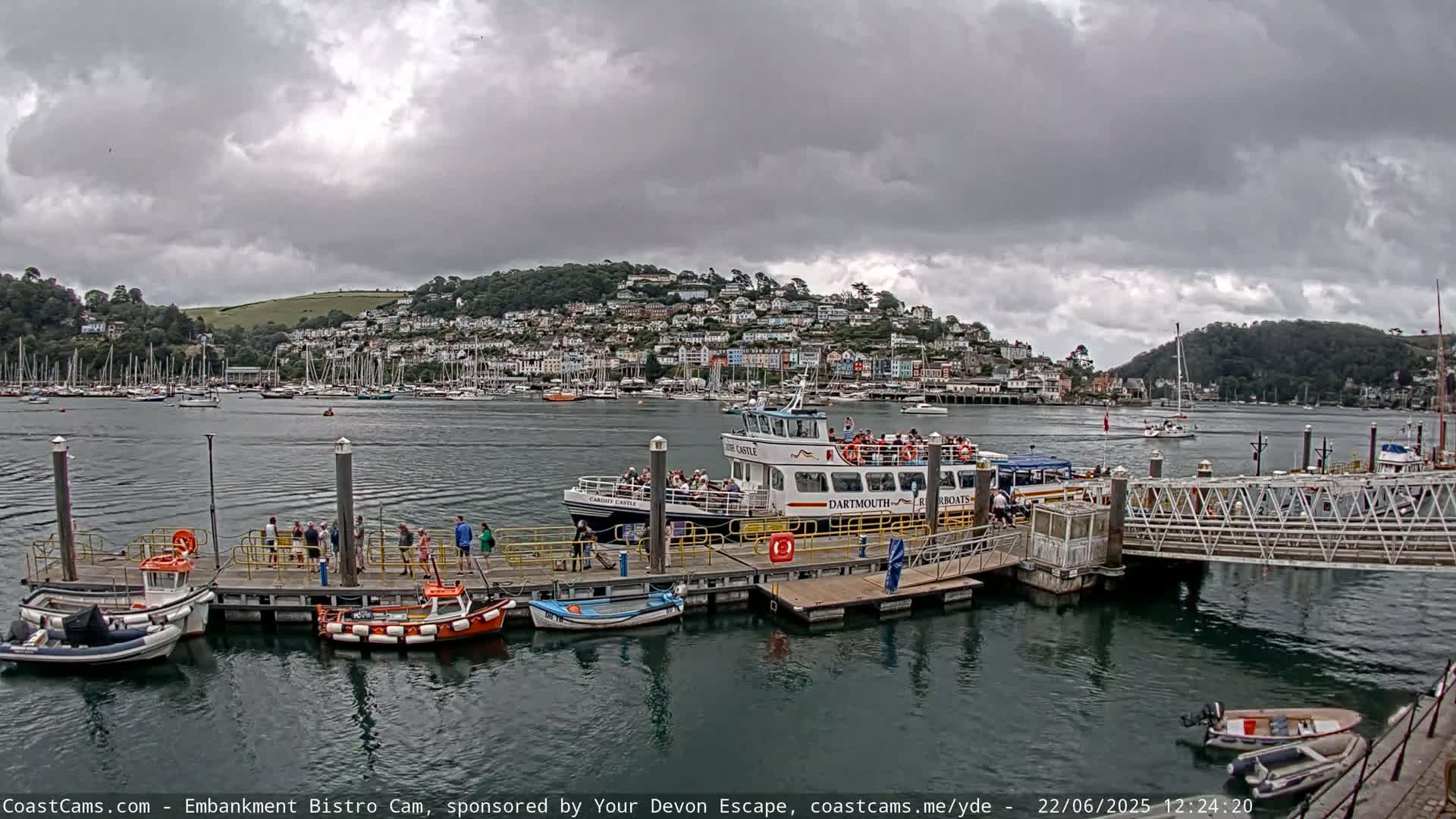 A passenger ferry is docked at a pier in a harbor filled with boats, under a cloudy sky.