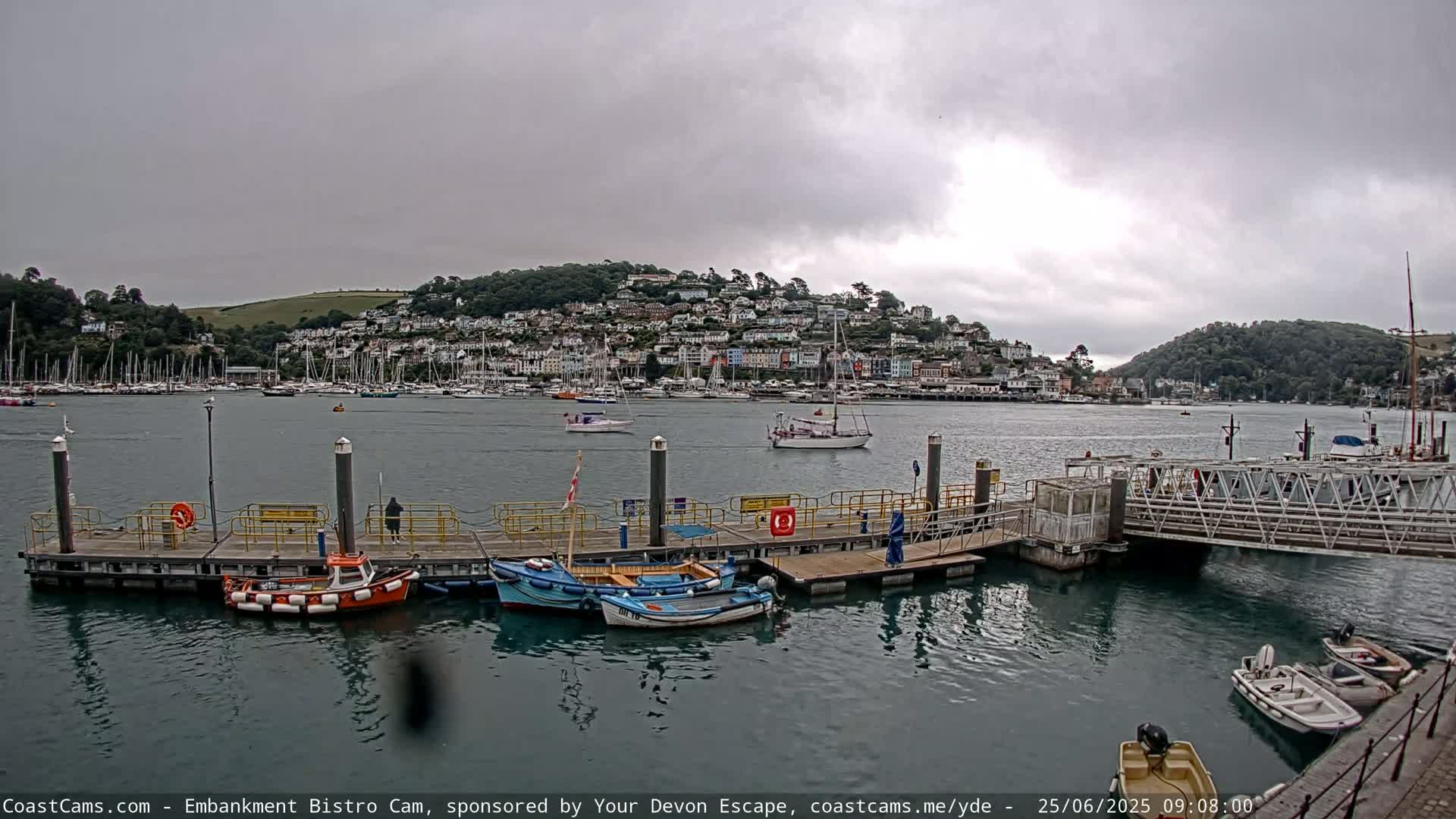 A harbor scene with overcast skies shows several small boats moored at a dock in front of a town nestled on a hillside.