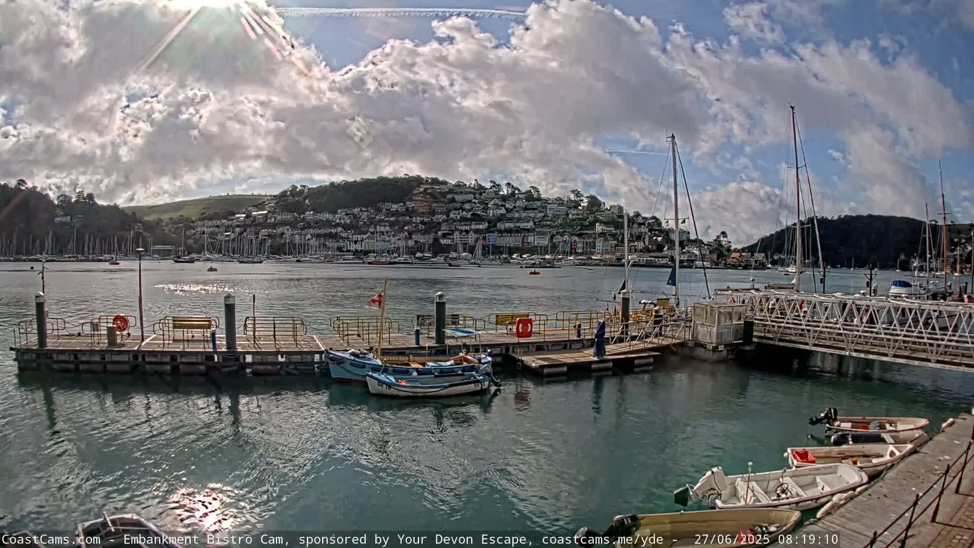A harbor with numerous boats moored at a dock, under a partly cloudy sky, with a hillside town visible in the background.