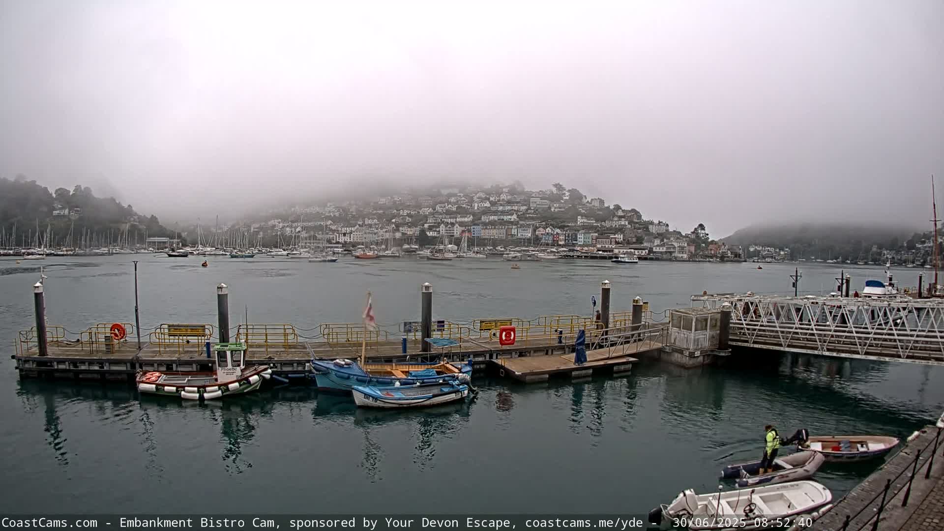 A foggy harbor scene shows a town nestled on a hillside overlooking a calm body of water with several small boats moored at a dock.