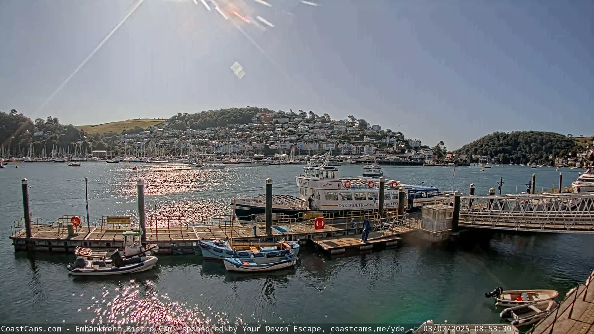 A sunny day at a harbor shows several boats docked at a pier, with a larger passenger ferry and a town nestled on a hillside in the background.