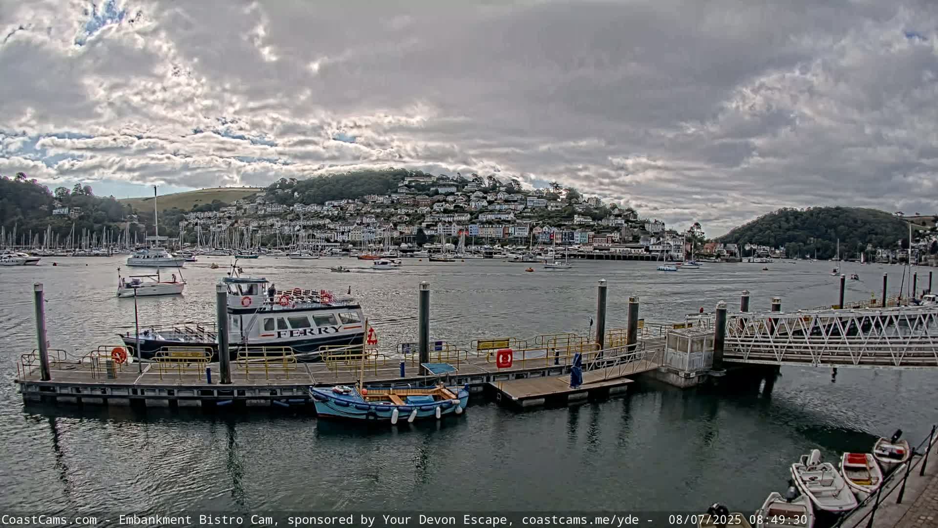 A ferry is docked at a pier in a harbor, under a cloudy sky, with numerous boats and a hillside town visible in the background.
