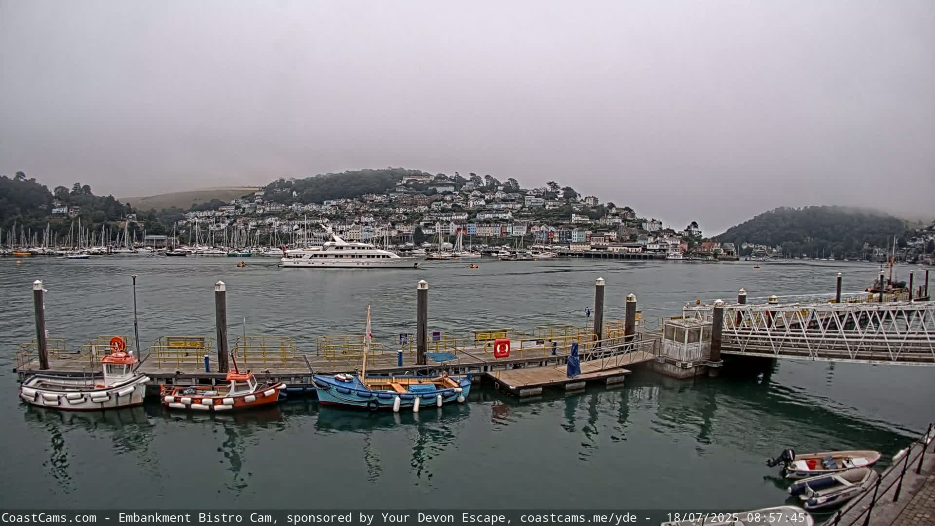 A harbor with several boats docked at a pier, a larger boat in the distance, and a town on a hill in the background under an overcast sky.