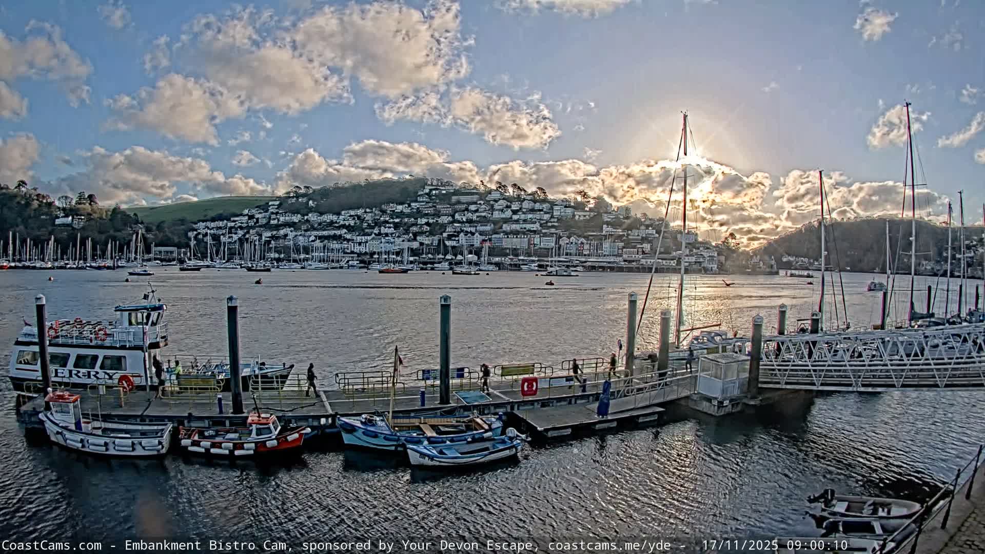 A harbor or river scene is depicted on a partly cloudy and sunny day, with a pier in the foreground featuring moored small boats and people walking, while numerous sailboats fill the water leading to a town nestled on a distant hillside under bright sunlight.