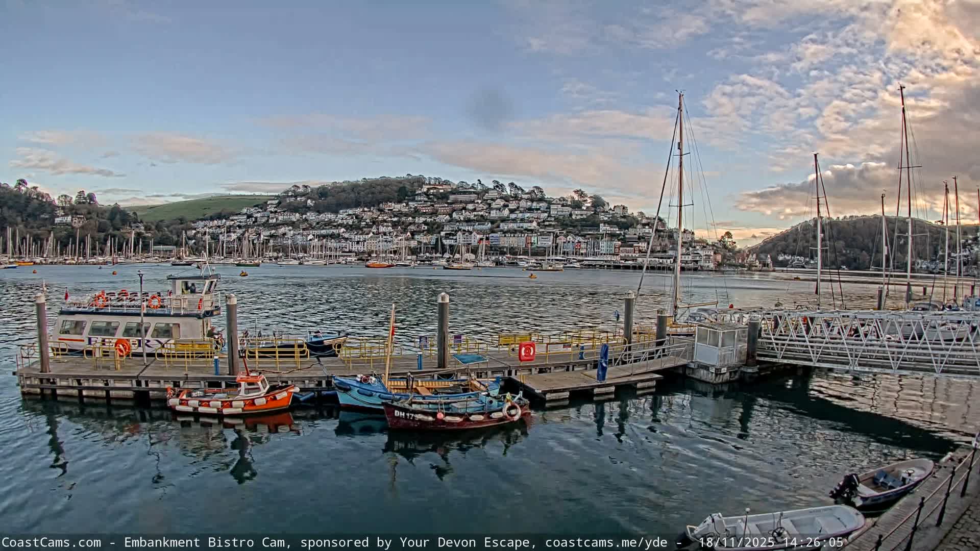 A harbor or river scene is depicted on a partly cloudy and sunny day, with a pier in the foreground featuring moored small boats and people walking, while numerous sailboats fill the water leading to a town nestled on a distant hillside under bright sunlight.