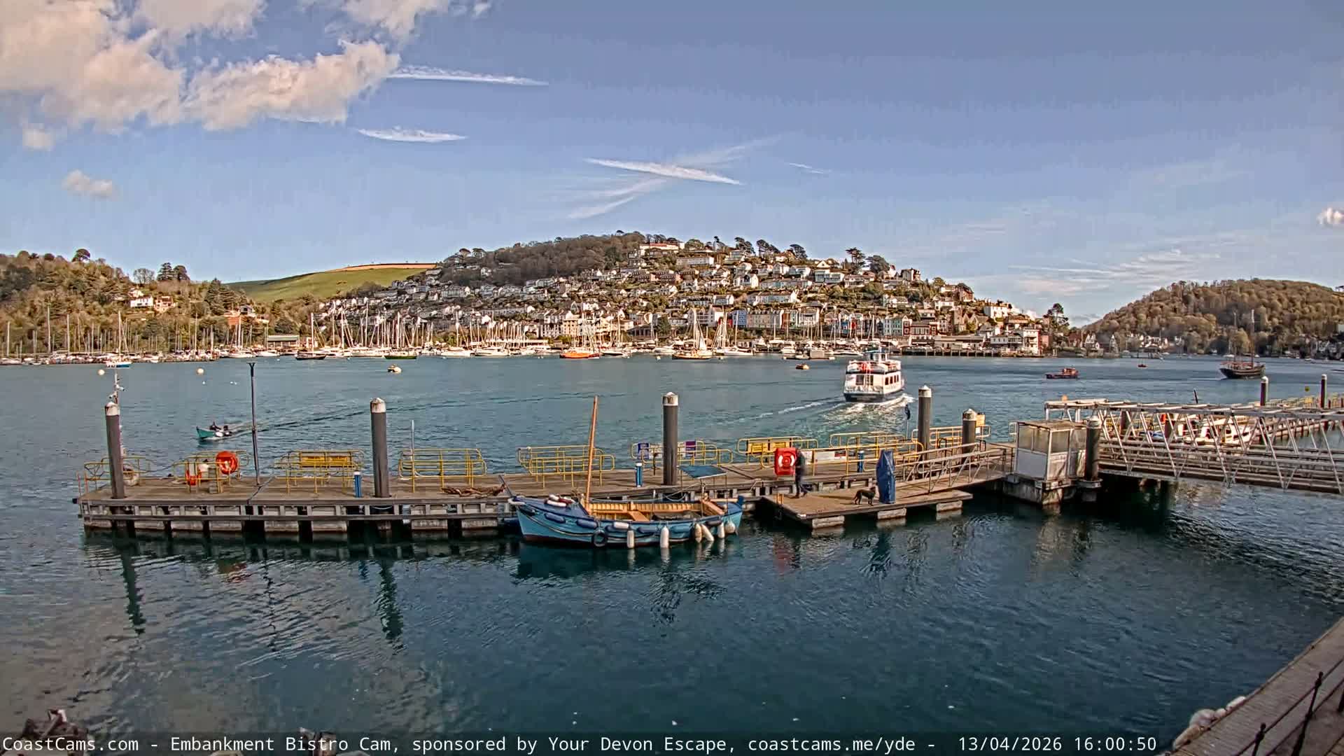 A harbor or river scene is depicted on a partly cloudy and sunny day, with a pier in the foreground featuring moored small boats and people walking, while numerous sailboats fill the water leading to a town nestled on a distant hillside under bright sunlight.