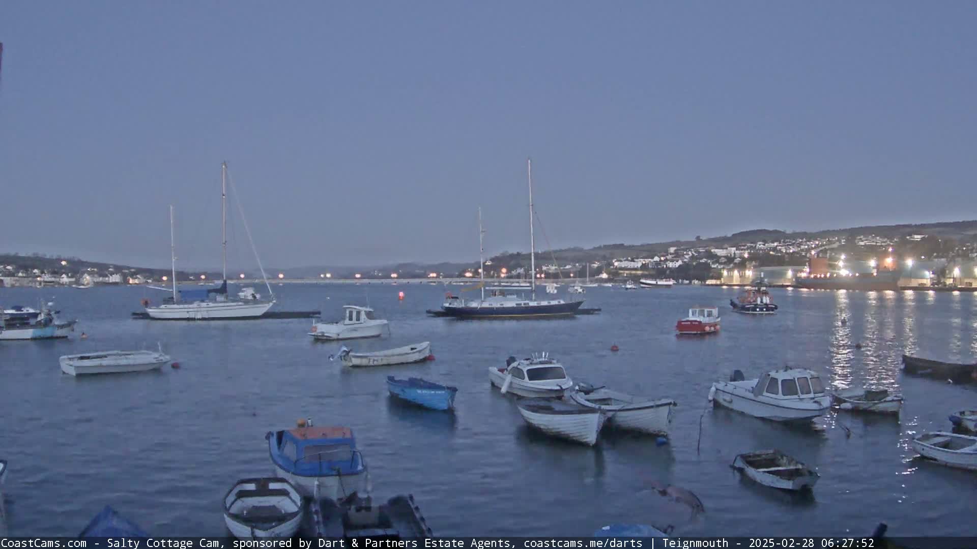 Numerous small boats and sailboats are moored in a calm harbor at twilight, with a town visible in the background.
