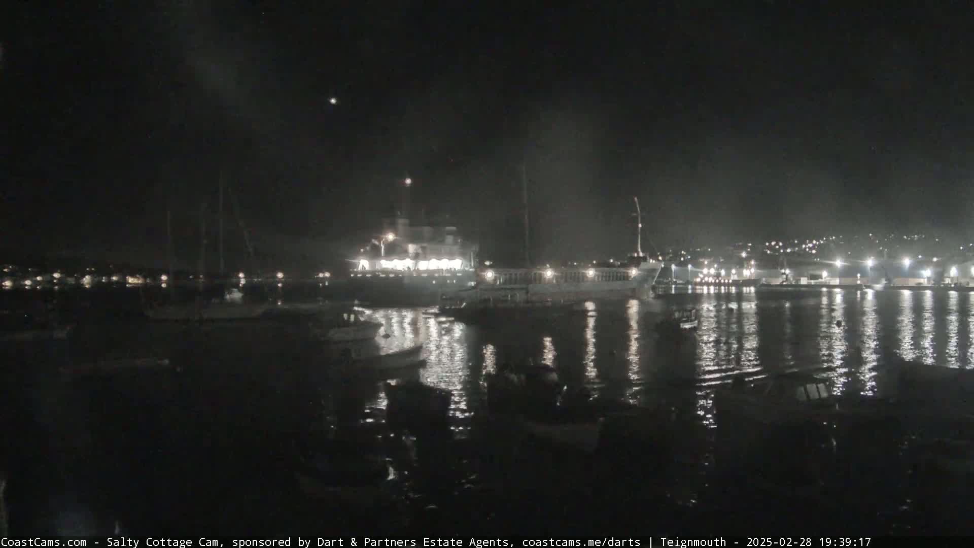 A nighttime harbor scene shows several boats moored in calm water under a dark, clear sky.
