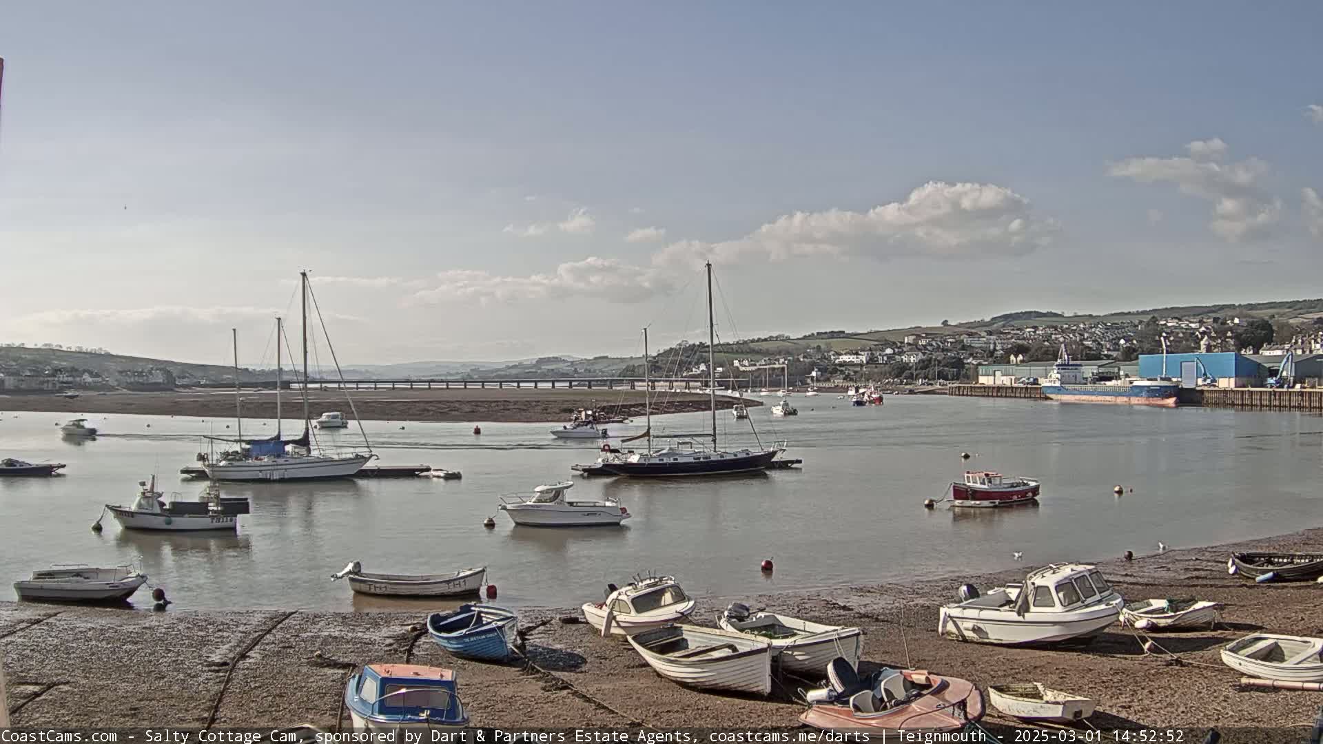 A harbor filled with sailboats and motorboats sits on a calm, partly cloudy day, with a few boats aground on a sandy shore.