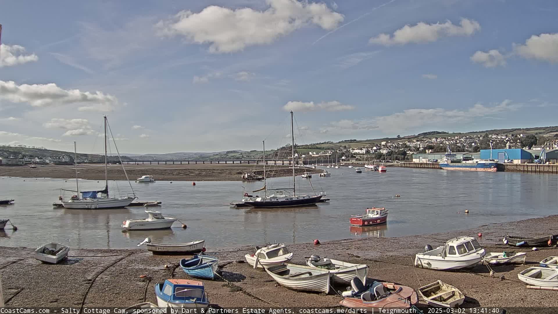A harbor scene on a partly sunny day shows numerous small boats on the mudflats and several sailboats and motorboats in the water, with a town and bridge visible in the distance.