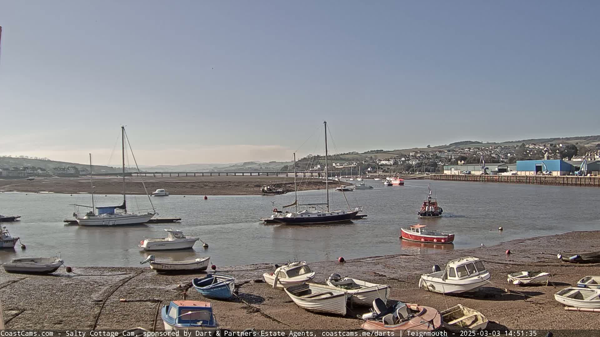 Numerous boats are moored in a calm harbor on a sunny day, with a low tide exposing a wide expanse of mudflats.