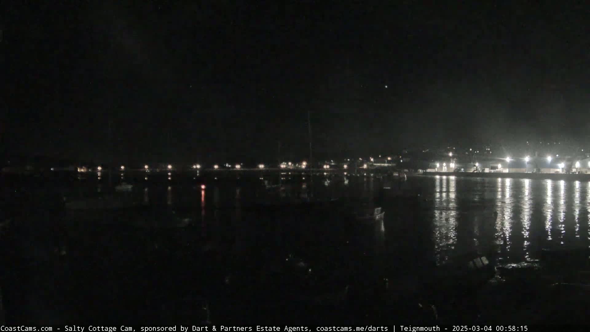 A nighttime view of a harbor with boats and lights reflecting on calm water under a clear, dark sky.