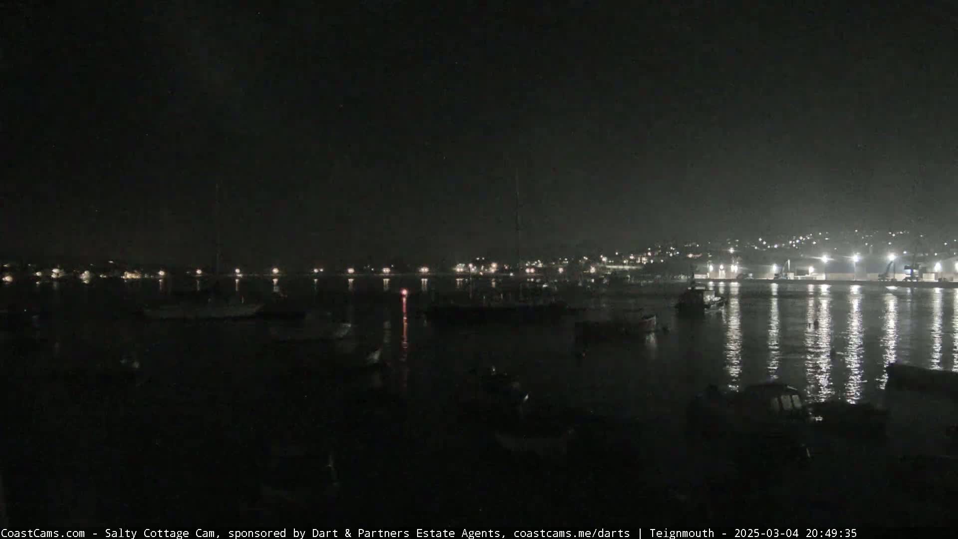 A nighttime view of a calm harbor with boats and a town's lights reflected on the water.