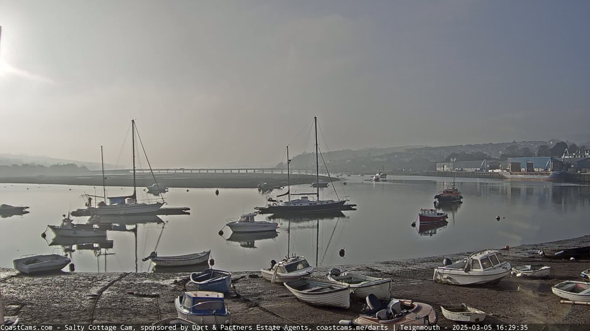 A hazy morning reveals a calm harbor with numerous sailboats and motorboats moored, some on land and others afloat, under a light sky.