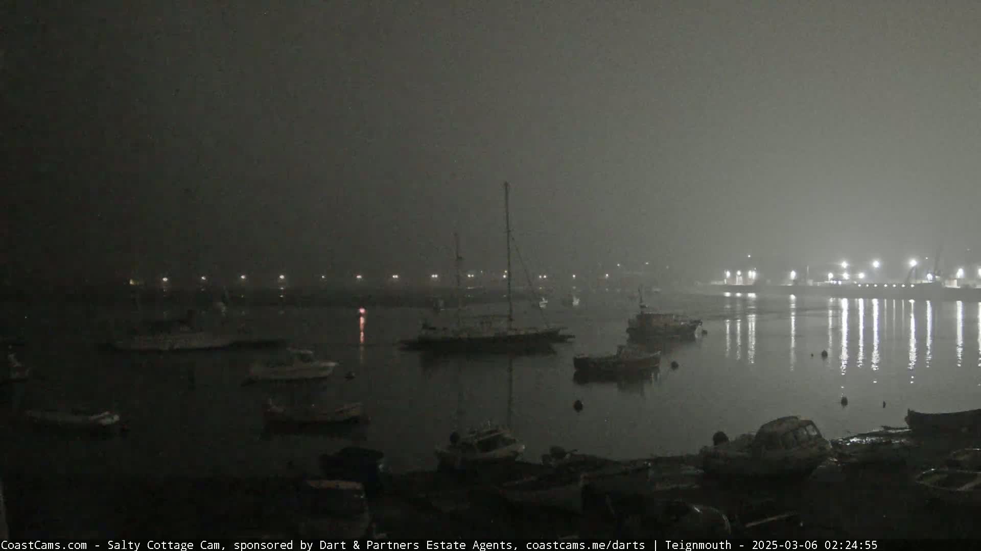 A foggy night scene shows numerous boats moored in a harbor, with lights faintly visible in the distance.