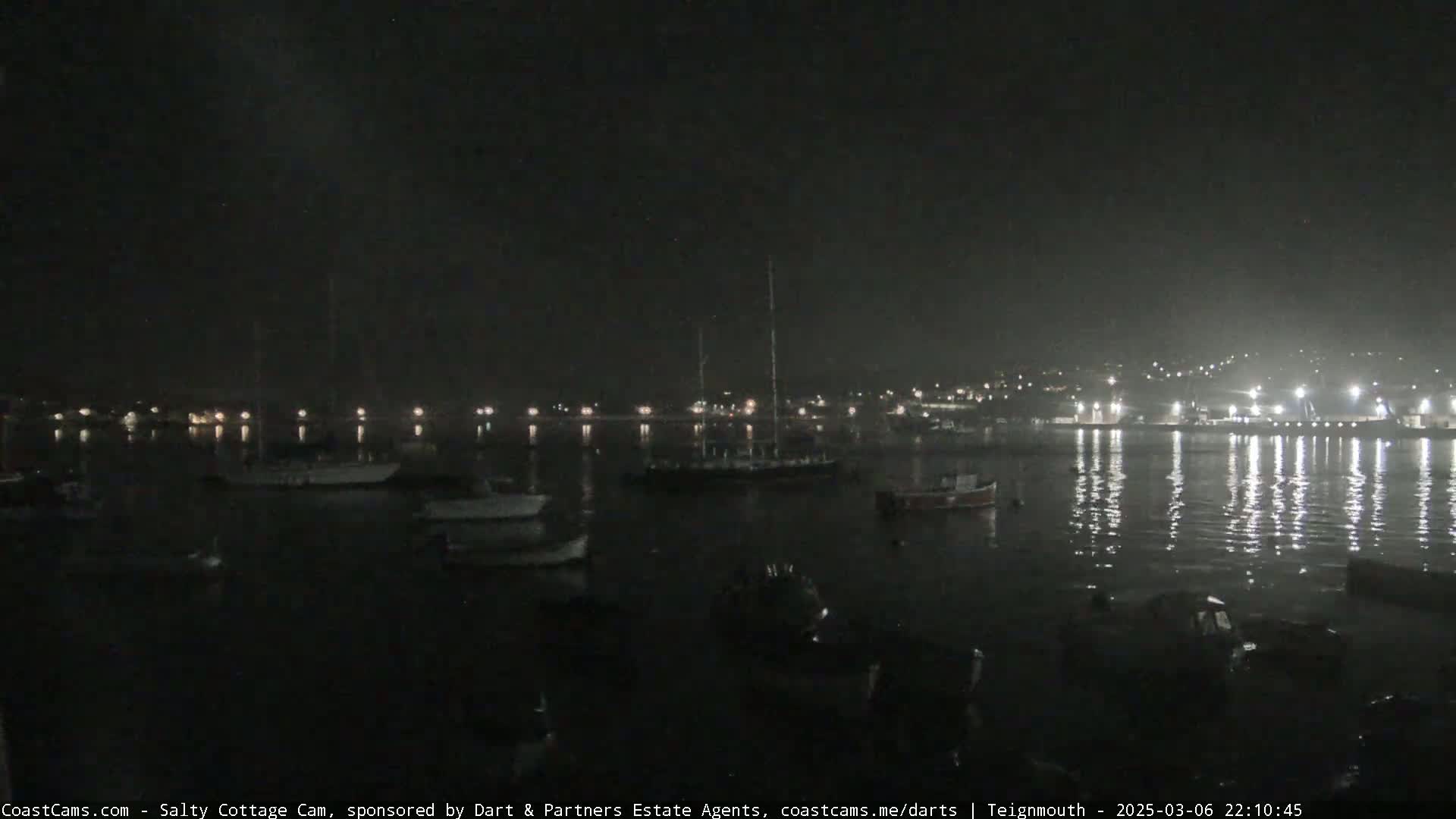 A nighttime harbor scene shows several boats moored in calm water, with lights reflecting on the water's surface and a town's lights visible in the distance.