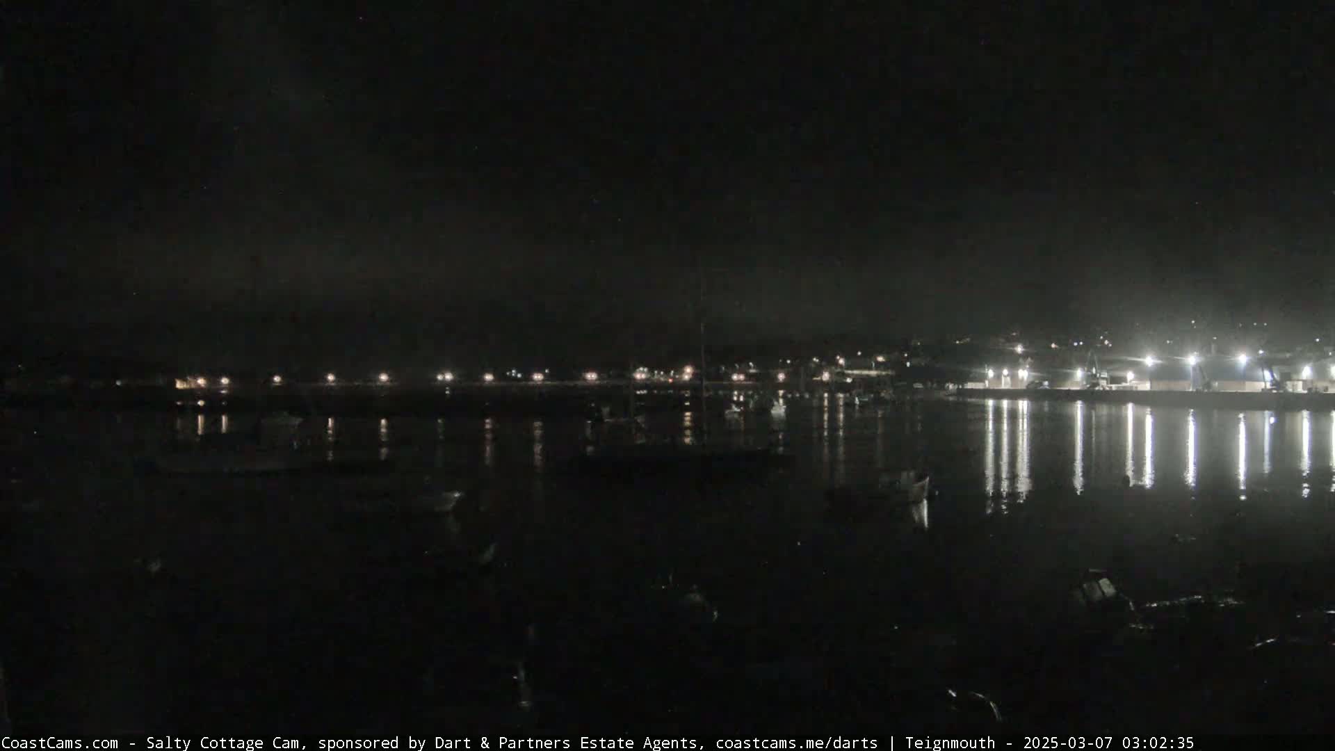 A nighttime view of a calm harbor with boats and lights reflecting on the water under a dark, clear sky.