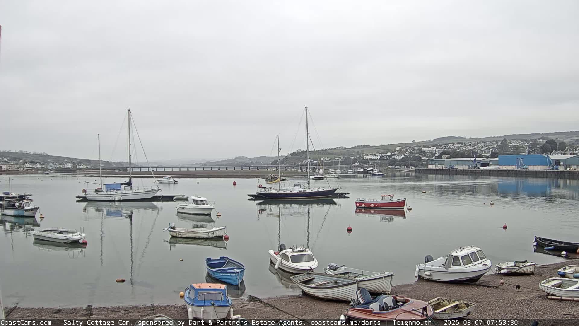 A calm, overcast day at a harbor shows many small boats and sailboats moored, with a town and hills visible in the background.