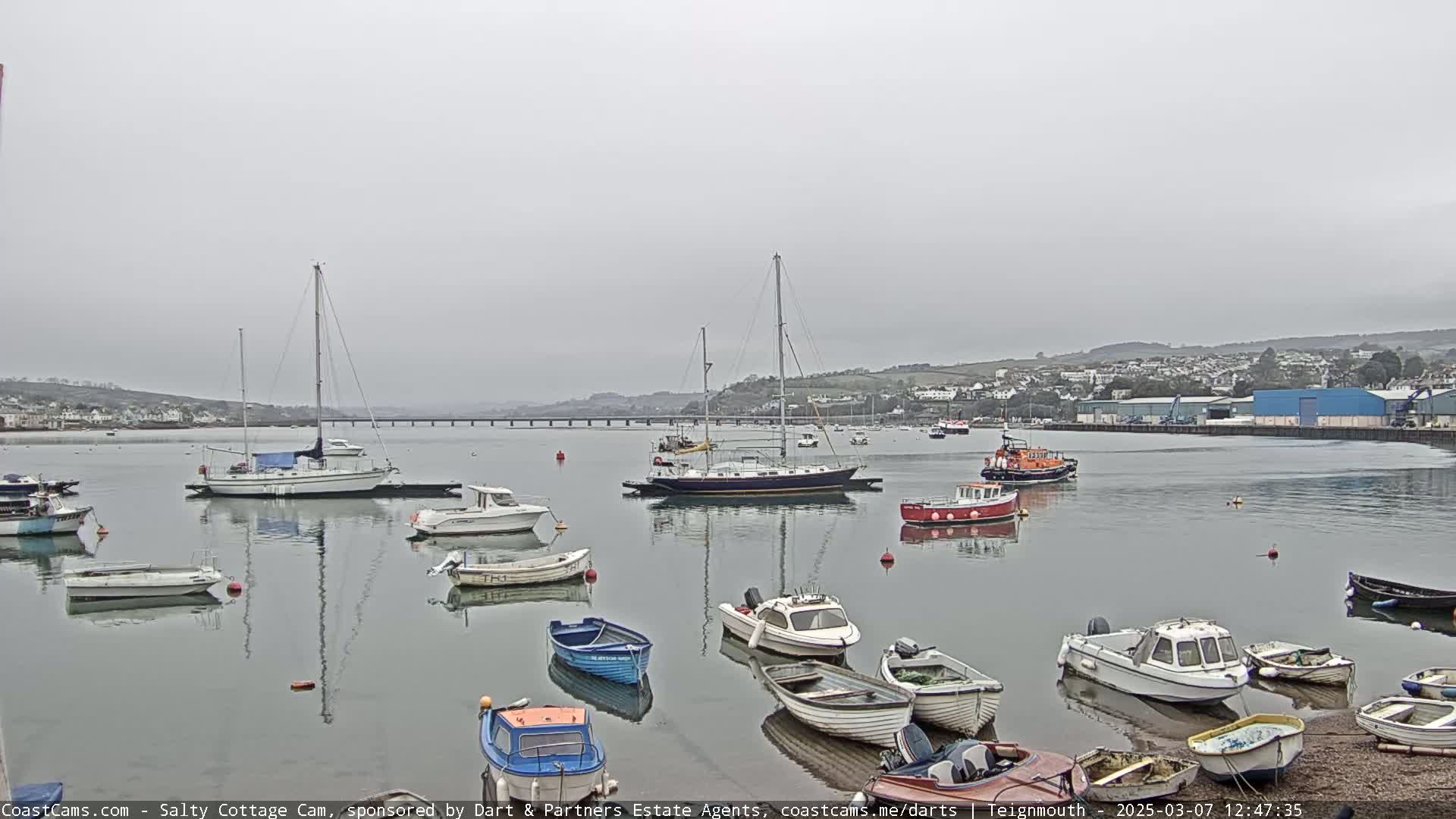A calm, overcast day reveals a harbor filled with various sailboats and motorboats, moored near a shoreline with a bridge visible in the distance.