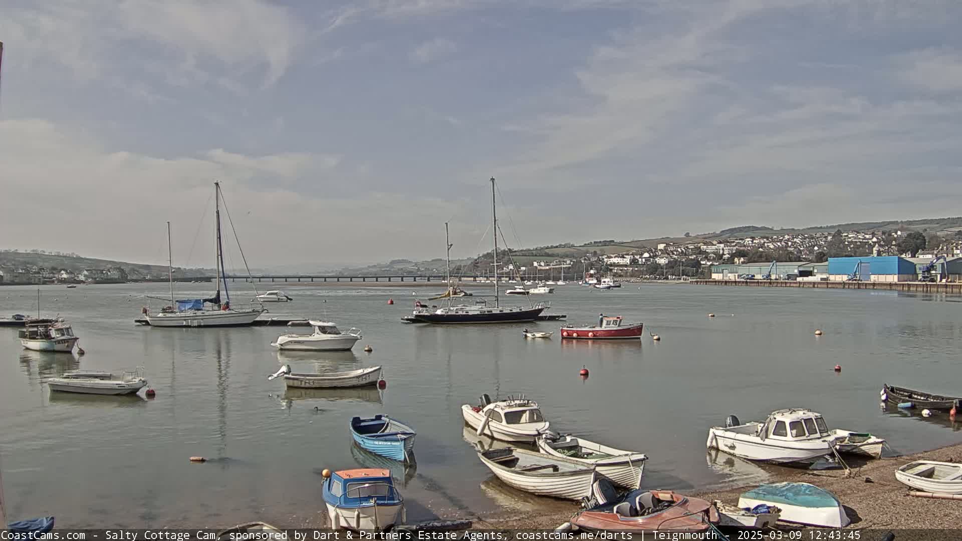 A calm, partly cloudy day shows many small motorboats and sailboats moored in a harbor, with a town and bridge visible in the distance.