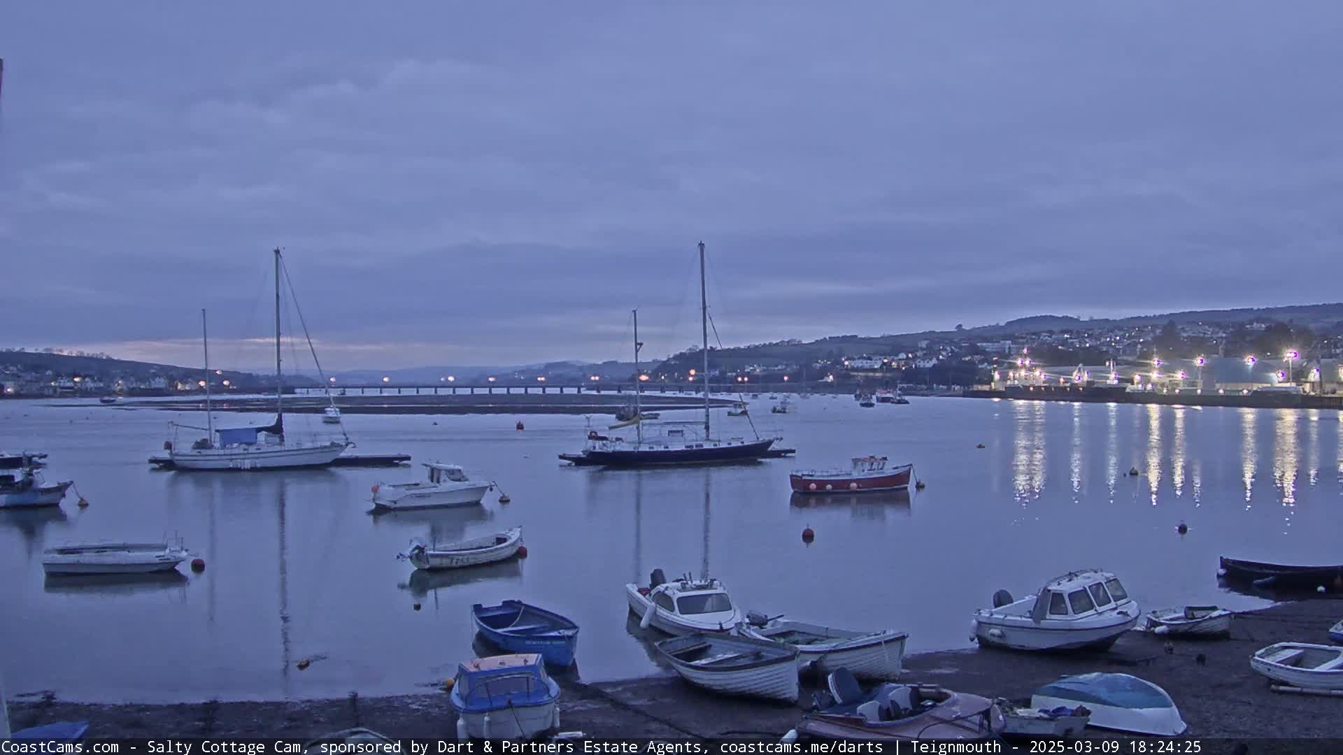Several sailboats and motorboats are moored in a calm harbor at dusk under a cloudy sky.
