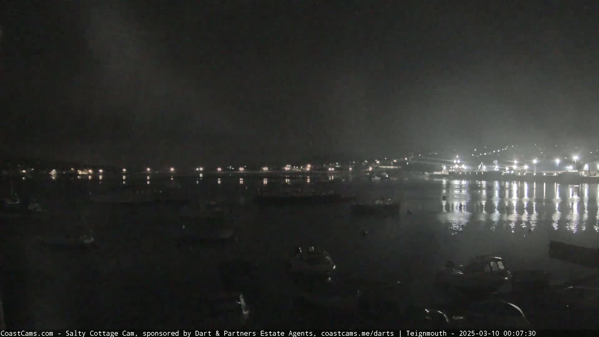 A nighttime view of a harbor with many boats and lights reflecting on the water under a dark, cloudy sky.