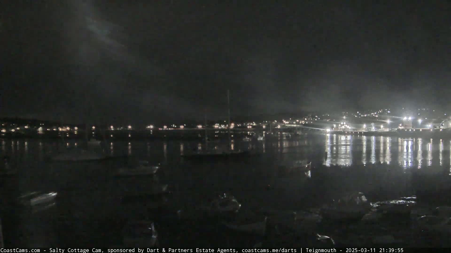 A nighttime view of a harbor with boats and a town's lights reflecting on calm water under a dark, cloudy sky.