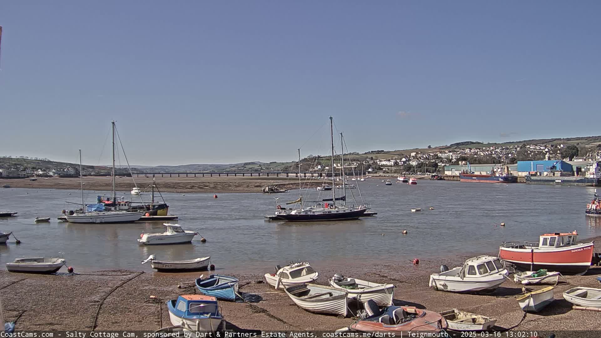 A harbor scene on a sunny day with numerous boats, some docked on a mudflat and others anchored in the calm water, a bridge in the distance, and a town visible on a hillside across the water.