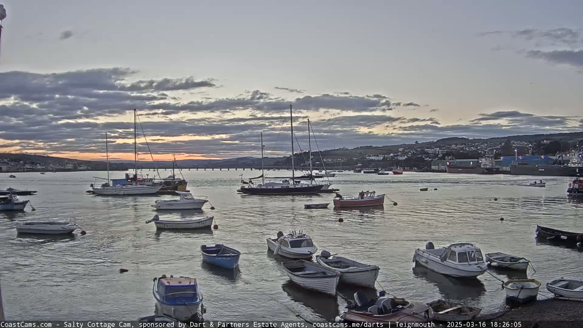 Numerous small boats and sailboats are moored in a calm harbor at sunset under a partly cloudy sky.