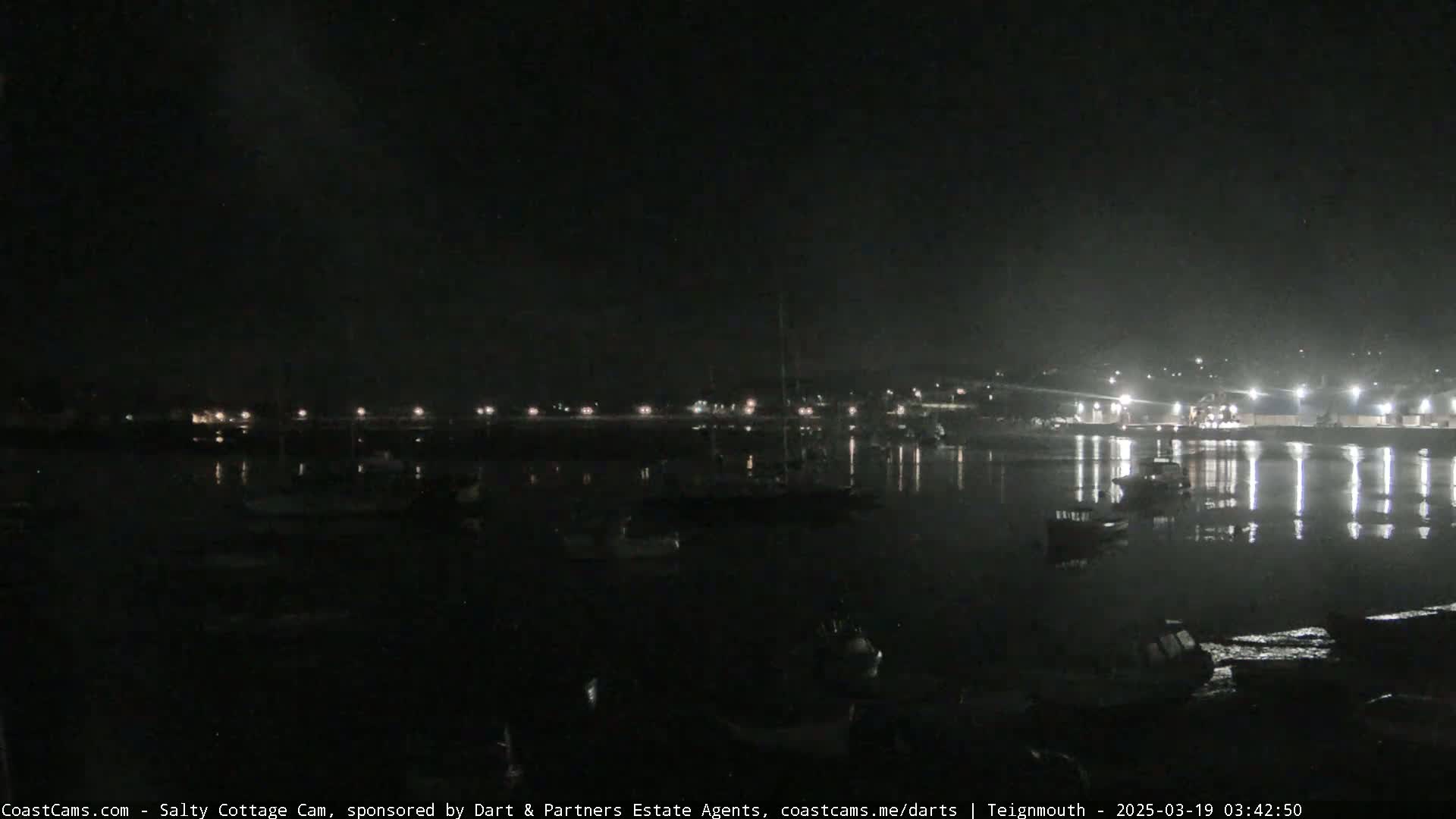 A nighttime view of a calm harbor with boats moored, reflecting lights from a distant shore under a dark sky.