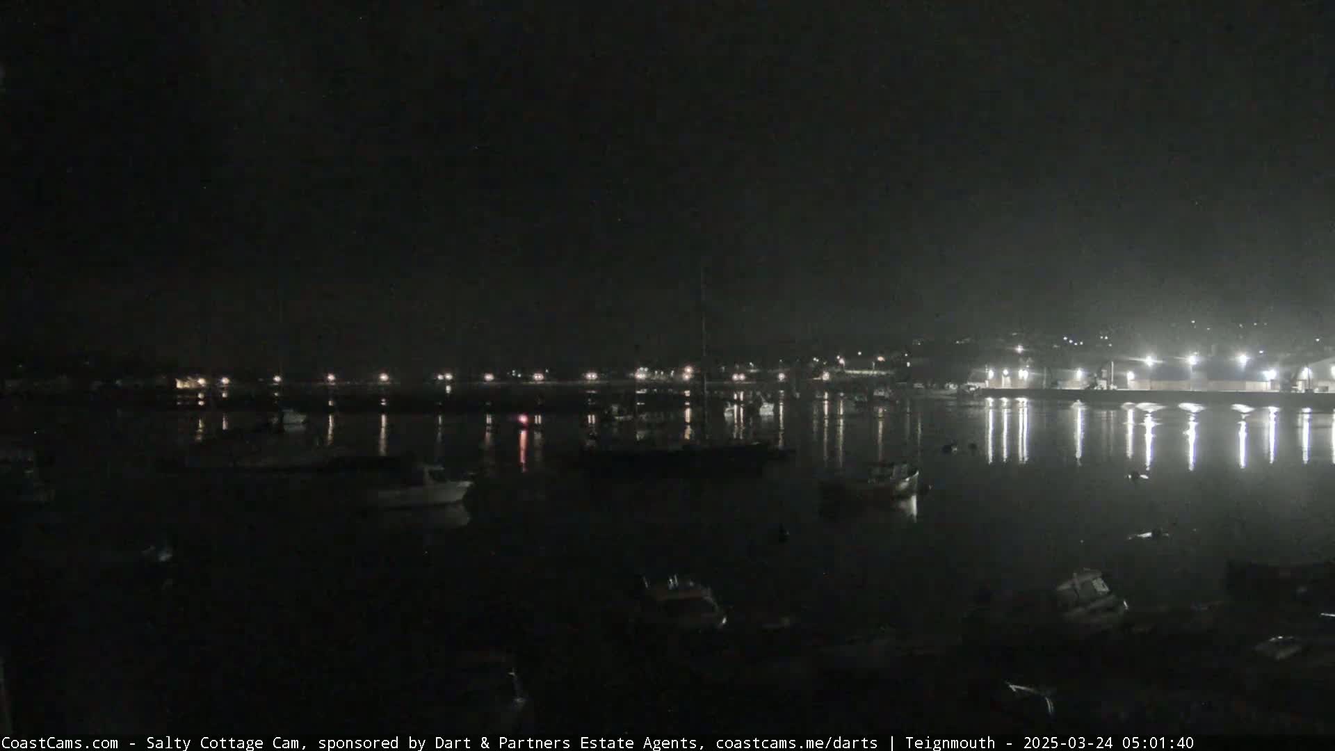 A nighttime view of a calm harbor with several boats moored, illuminated by lights along the waterfront.