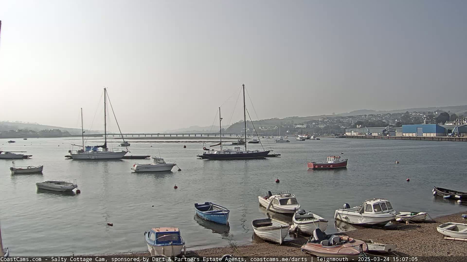 Numerous boats are moored in a calm harbor on a hazy day, with a bridge visible in the distance.