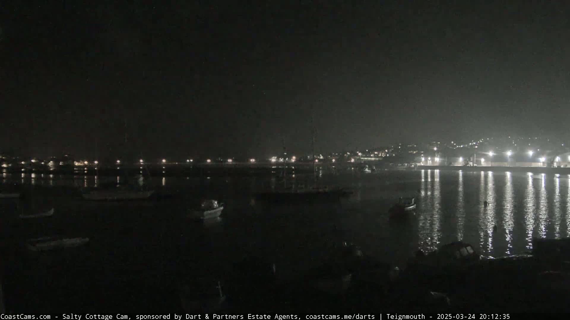 A nighttime view of a harbor with several boats moored, illuminated by lights from the shore.