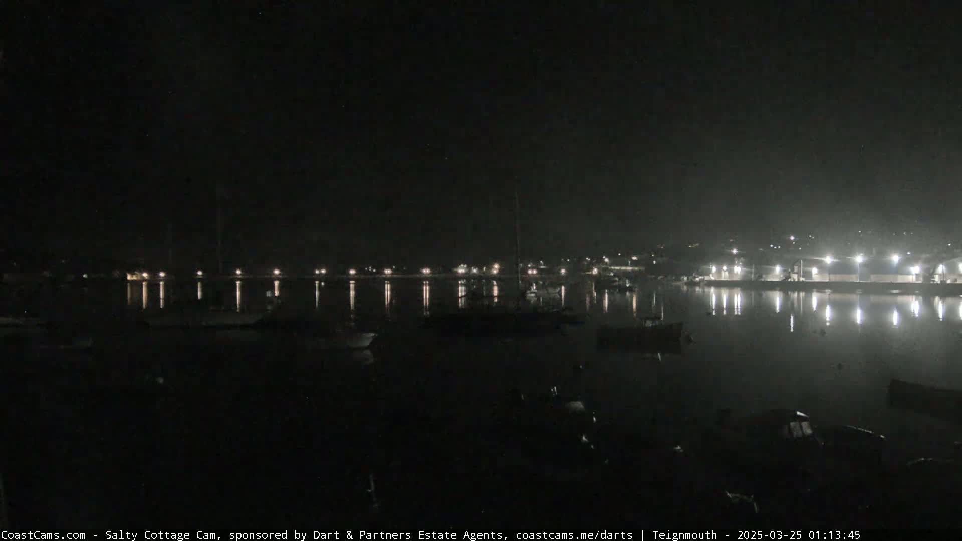A dark nighttime harbor scene shows a calm body of water reflecting the lights from buildings and boats on the shore.