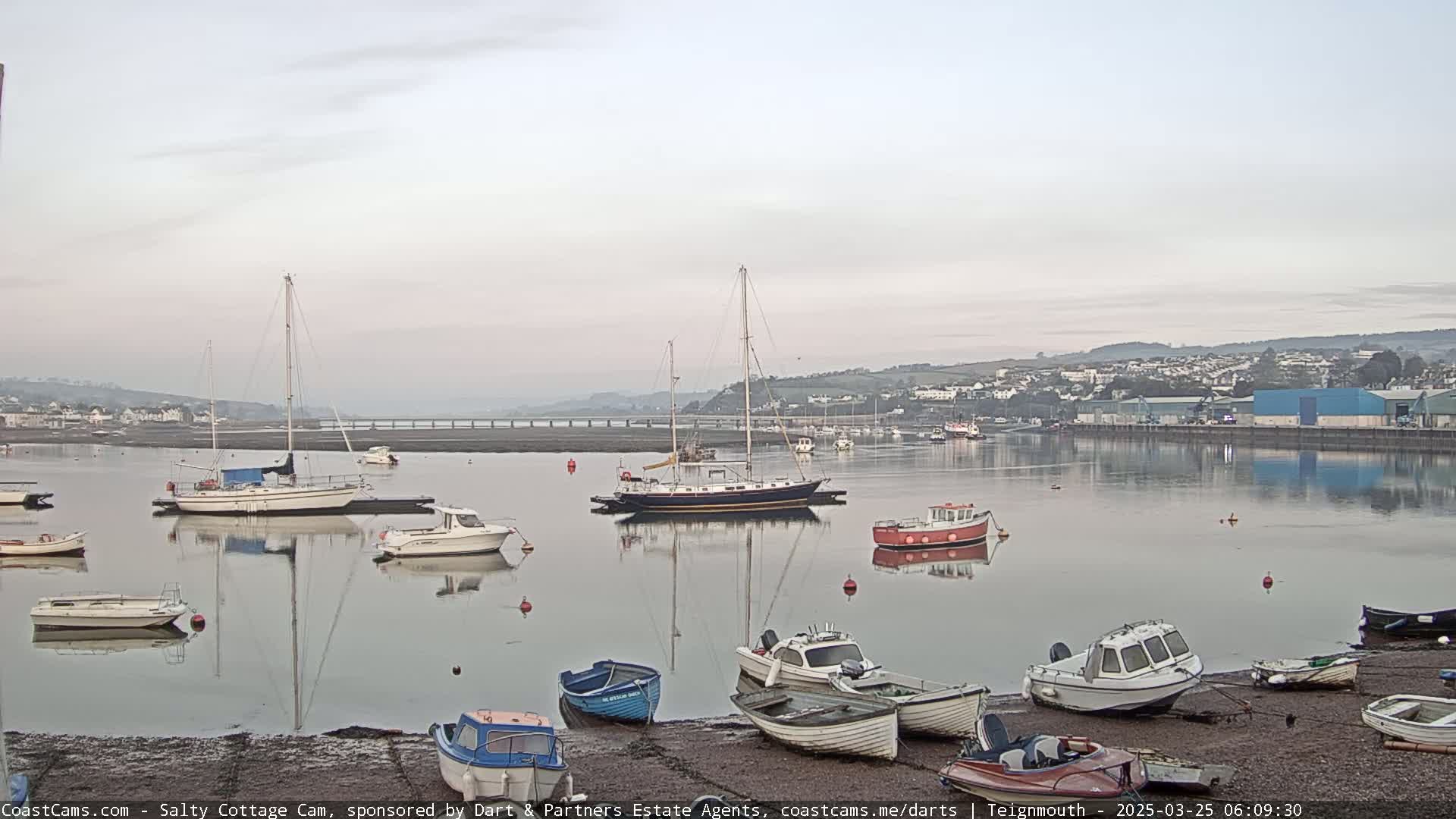 Several sailboats and motorboats are moored in a calm harbor at dawn, with a town and bridge visible in the background.