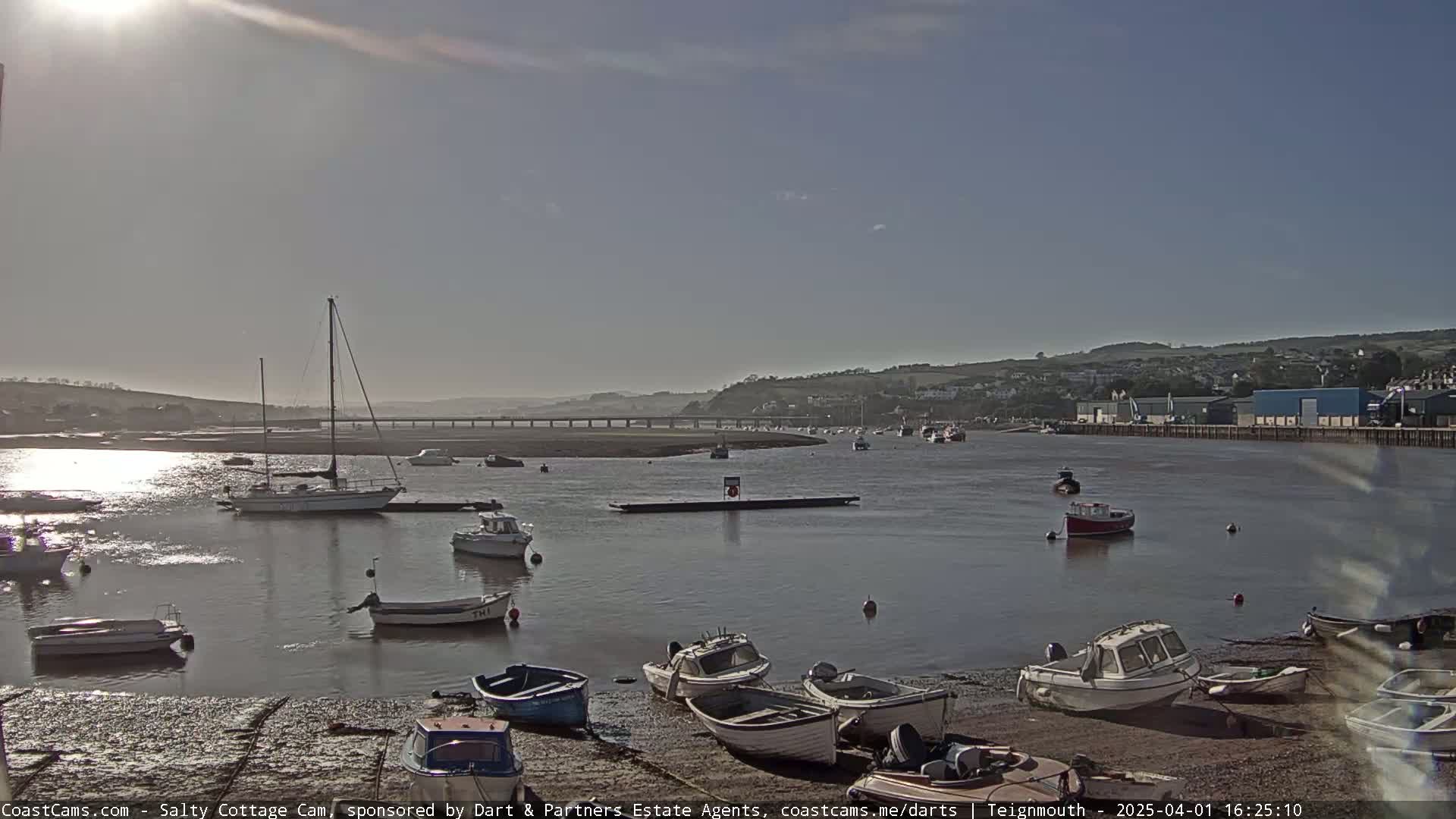 A sunny day at a harbor shows many small boats docked along a shoreline with a bridge and distant hills in the background.