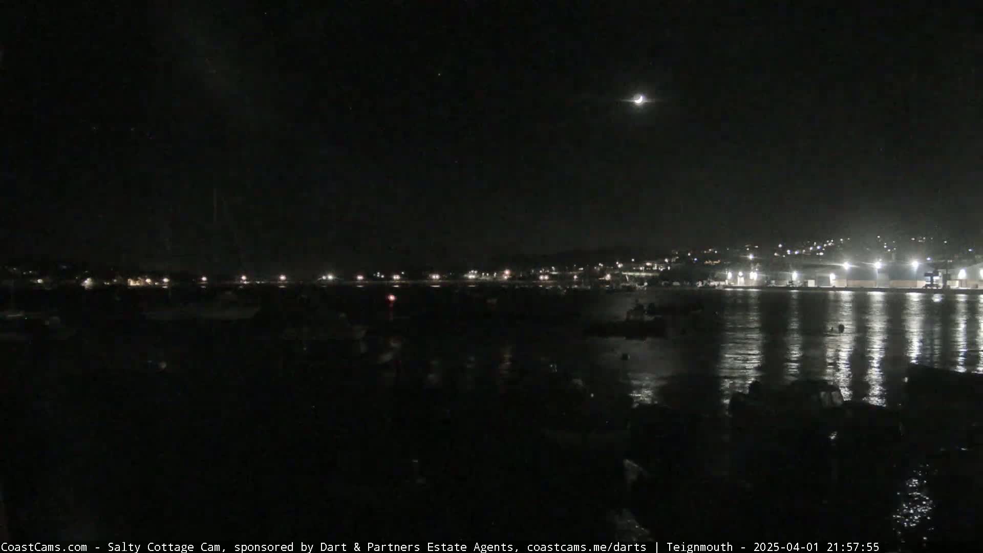 A crescent moon hangs in a dark, clear night sky over a calm body of water reflecting the lights of a distant town.