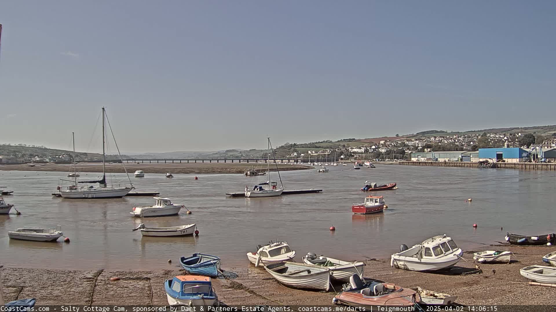 Numerous small boats are moored in a calm, brown-water estuary on a sunny day, with a town visible in the distance across the water.