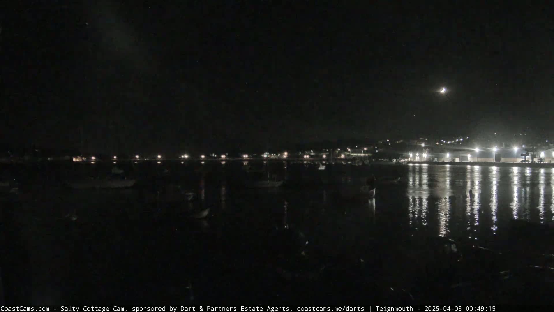A crescent moon is visible in the night sky over a dark harbor with several boats, and lights from buildings are reflected in the calm water.