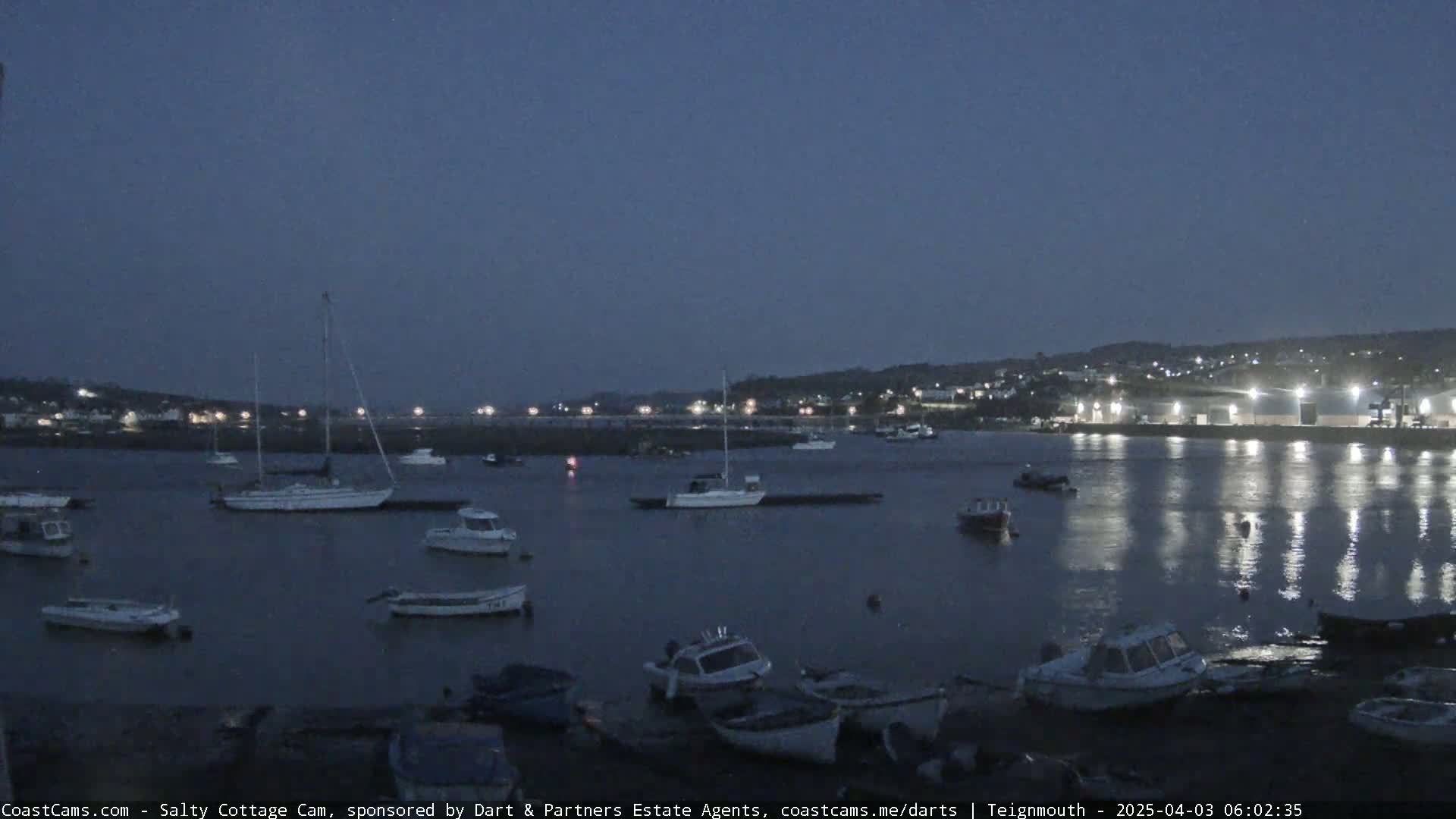 Many small boats are moored in a calm harbor at twilight, with a town visible in the background.