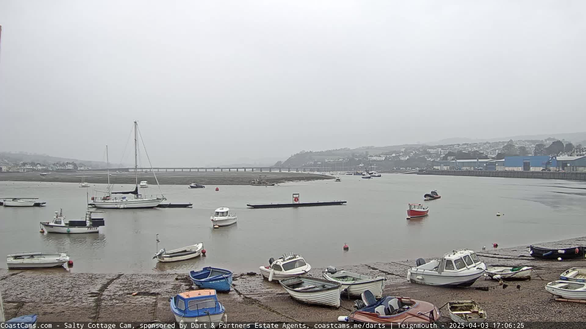 Numerous small boats are moored on a muddy shoreline next to calm water under an overcast sky, with a bridge and distant town visible in the background.