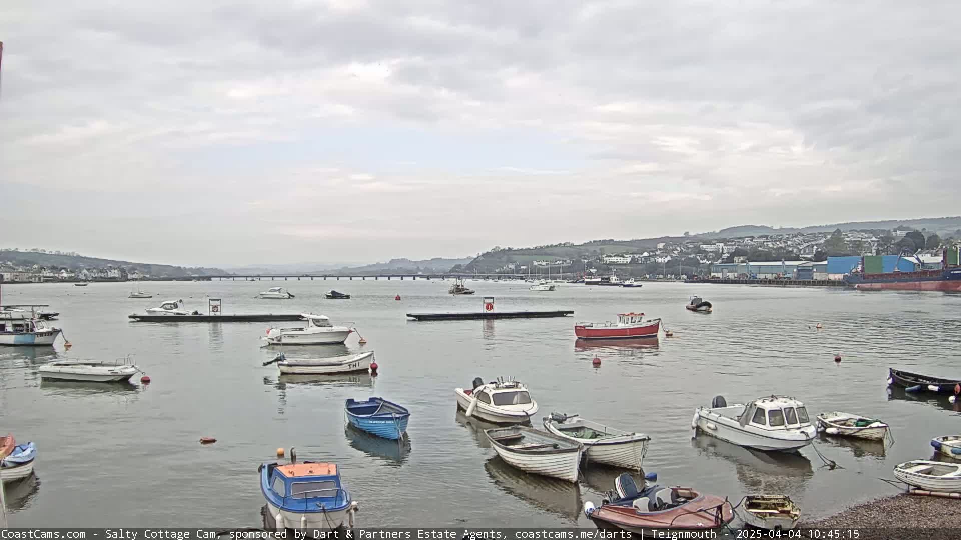Numerous small boats are moored in a calm harbor under an overcast sky, with a distant town and bridge visible across the water.