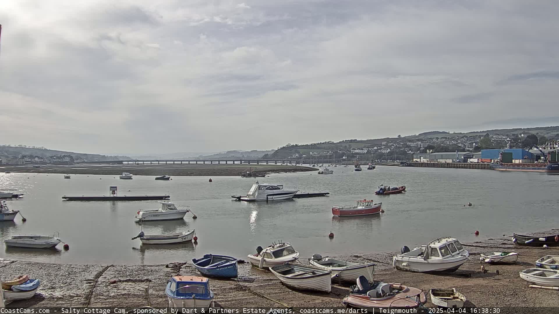Numerous small boats are moored in a calm harbor under a mostly cloudy sky, with a bridge and distant buildings visible across the water.