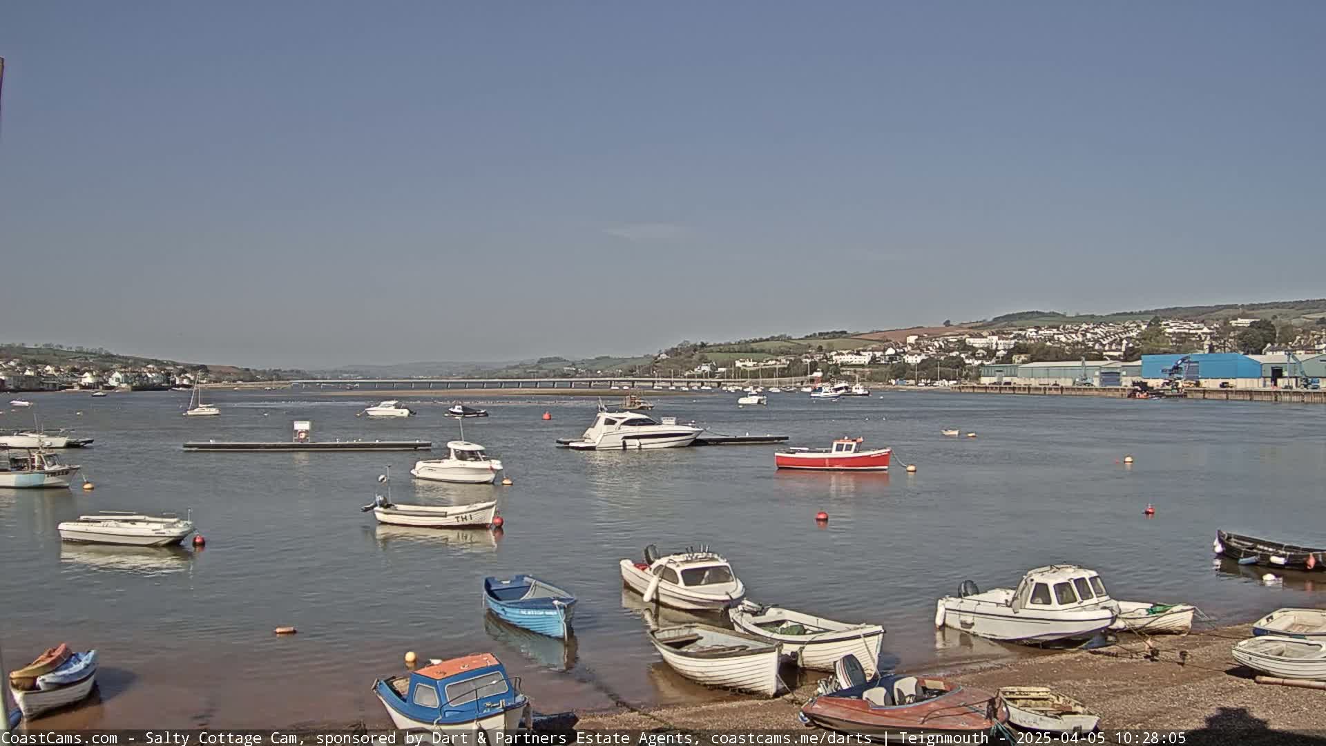 A calm, sunny day shows numerous small boats moored in a harbor, with a bridge and distant buildings visible across the water.