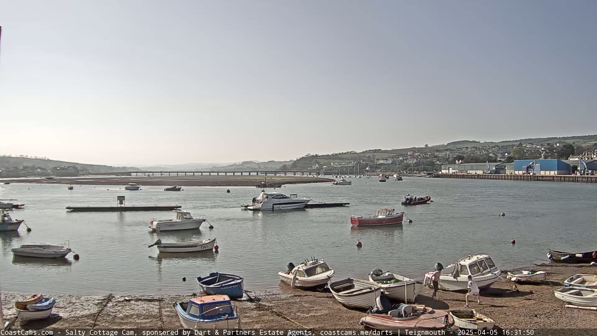 A calm, sunny day reveals a harbor filled with numerous small boats and a few larger vessels, nestled against a shoreline with hills in the background.