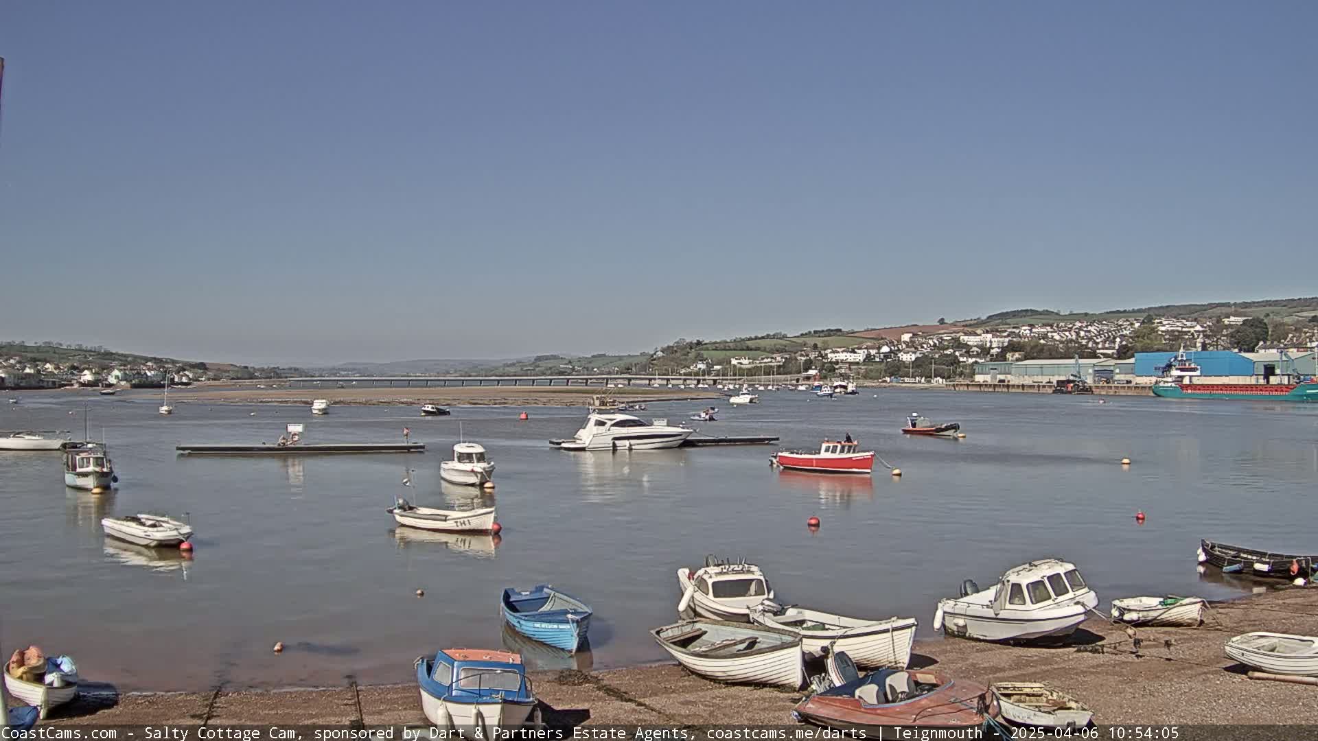 Numerous small boats are moored in a calm harbor on a sunny day, with a town and a bridge visible in the distance.